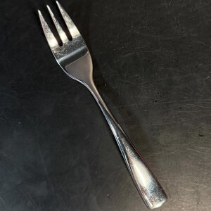 Puede incluir: Un tenedor de acero inoxidable con tres dientes y un mango liso y pulido. El utensilio tiene un acabado metálico reflectante y un diseño cónico, con un cuello curvo que conecta la cabeza con la base sólida y ligeramente ensanchada.