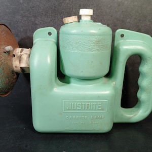 May include: Vintage green Justrite carbide lamp with a handle and a reflector. The lamp has a cylindrical fuel container and a rusty reflector. The text "JUSTIRITE CARBIDE LAMP" is visible on the front of the lamp.