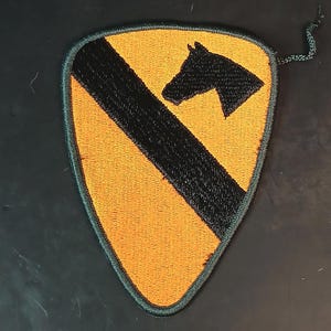 May include: Embroidered patch featuring a shield shape with a black horse head and a black diagonal stripe on a yellow background. The patch has a green border and a small string attached.