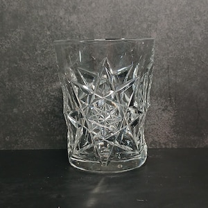 May include: A clear, faceted glass tumbler with a starburst pattern. The glass has a thick base and a straight, slightly flared rim. The intricate design catches the light, creating a sparkling effect. The glass is empty and sits on a dark surface.
