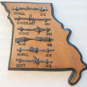Könnte beinhalten: Holztafel in Form des Bundesstaates Missouri, mit verschiedenen Stacheldrahtdesigns. Jeder Drahttyp ist mit einem Namen und Jahr gekennzeichnet, wie "Crandal 1879" und "Sunderland 1884". Die Tafel hat einen dunklen Rand.