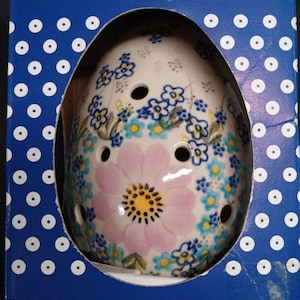 Pode incluir: Um objeto decorativo em cerâmica em forma de ovo com um design floral, apresentado numa caixa azul e branca com bolinhas. O ovo apresenta uma grande flor rosa e detalhes florais azuis. A caixa diz "HANDMADE IN POLAND".