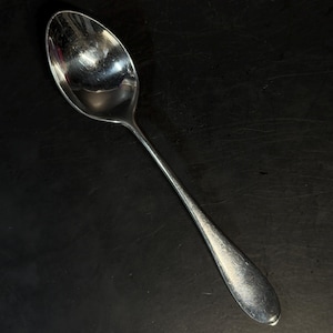 May include: A polished silver-toned metal spoon with an oval-shaped bowl and a smooth, slightly tapered handle. The surface of the spoon exhibits a reflective finish with subtle signs of wear, including fine scratches visible across the metal.