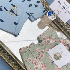 Op de afbeelding: Een lichtblauwe babyromper met een dinosauruspatroon, gecombineerd met een gouden hangerketting met een dinosaurus silhouet. Een bijpassende bloemenromper en zilveren hangerketting zijn ook aanwezig.