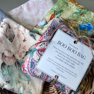 Puede incluir: Una colección de bolsas Boo Boo hechas a mano en una cesta de mimbre. Las bolsas presentan varias telas florales y estampadas en rosa, verde y azul. Una etiqueta atada con hilo dice "BOO BOO BAG" con instrucciones de uso.