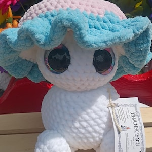 May include: A plush mushroom toy with a white body and a cap featuring a light pink top and a ruffled light blue edge. The toy has large, dark, reflective eyes and a white tag attached with twine that reads THANK YOU and includes care instructions.