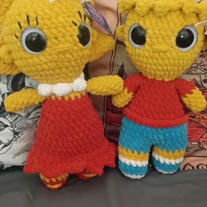 May include: Two handmade crochet dolls. One doll wears a red dress with a white collar. The other doll wears a red shirt and blue shorts with yellow and white stripes. Both dolls have yellow heads, large black eyes, and small arms.