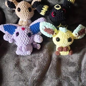 Peut inclure: Quatre peluches faites à la main en crochet, représentant des créatures de dessins animés. Les peluches sont de couleurs variées : beige, noir, violet et jaune, avec des détails comme des oreilles et des yeux. Elles sont posées sur une surface grise texturée.