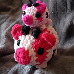 May include: A handmade crochet cow plushie in white, pink, and black. The cow has large black eyes and pink spots. The plushie is sitting on a gray, textured surface.