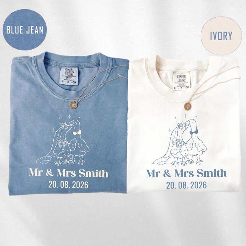 May include: Two t-shirts, one blue and one ivory, are displayed. Each shirt features a wedding-themed graphic with the text "Mr & Mrs Smith" and the date "20.08.2026". Both shirts have a necklace.