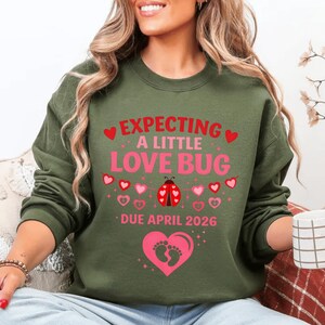 Op de afbeelding: Olijfgroene sweatshirt met de tekst "EXPECTING A LITTLE LOVE BUG" in roze en rood. Een lieveheersbeestjesafbeelding is gecentreerd onder de tekst, met een roze hart- en voetafdrukafbeelding onderaan. De tekst "DUE APRIL 2026" is ook opgenomen.