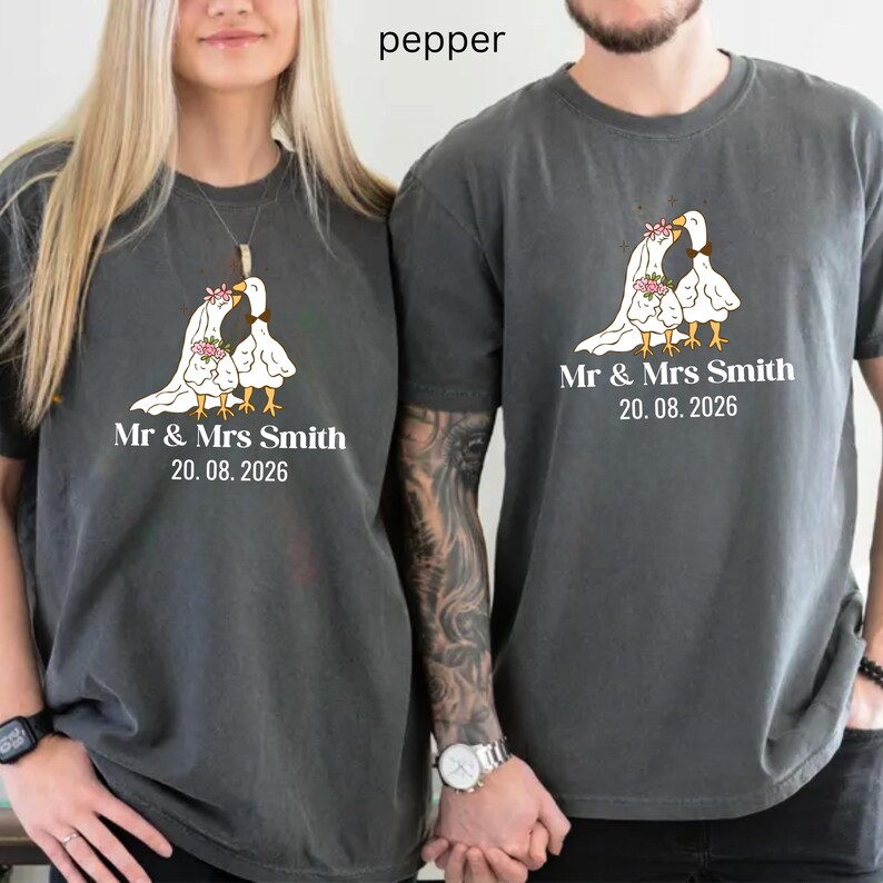 May include: Two gray t-shirts with a wedding design. The design features two white geese with floral accents, and the text "Mr & Mrs Smith 20.08.2026". The shirts are made of a soft material.