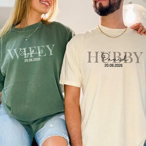 May include: Two matching shirts, one green and one cream. The green shirt says "Wifey" with a name and date. The cream shirt says "Hubby" with a name and date. Both shirts are casual and comfortable.