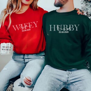 May include: A red crewneck sweatshirt with the word "Wifey", a name, and a date printed in white. A dark green crewneck sweatshirt with the word "Hubby", a name, and a date printed in white. Both sweatshirts are worn by a couple.