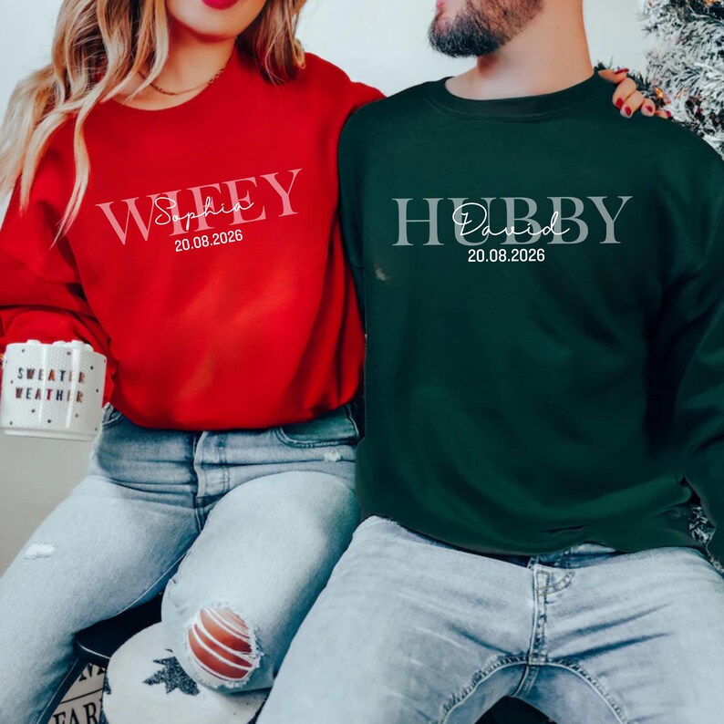 May include: A red crewneck sweatshirt with the word "Wifey", a name, and a date printed in white. A dark green crewneck sweatshirt with the word "Hubby", a name, and a date printed in white. Both sweatshirts are worn by a couple.