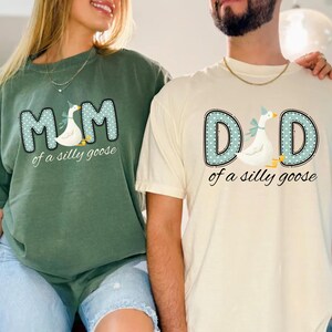 May include: Two t-shirts, one green and one cream-colored, feature the text "MOM" and "DAD" in a dotted blue font, each with a goose illustration wearing a party hat. The phrase "of a silly goose" is below the text.