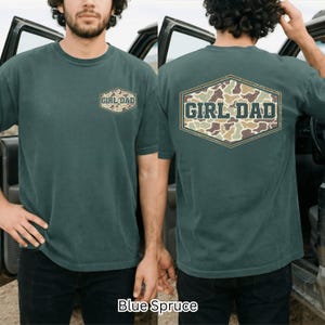 May include: Blue Spruce short-sleeve t-shirt with "GIRL DAD" text in a camouflage design. The text is on the front and back of the shirt. The shirt is a solid color with a crew neck.