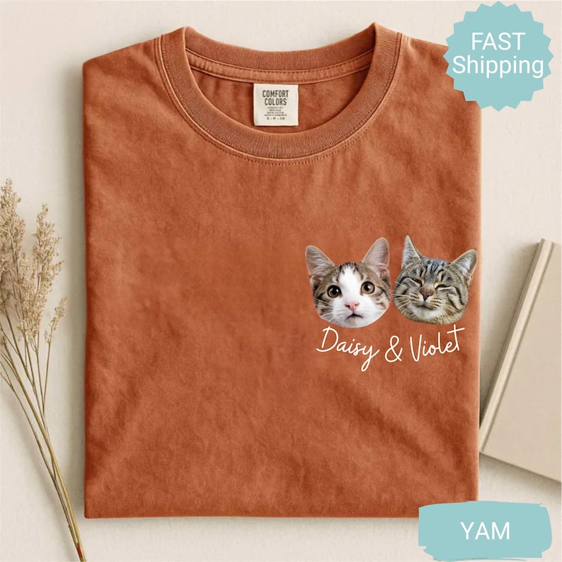 May include: A rust-colored t-shirt featuring two cat faces with the names "Daisy & Violet" printed below. The shirt has a crew neck and a small tag on the upper left. A "Fast Shipping" sticker is visible in the upper right corner.