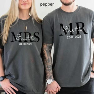 May include: Two pepper-colored t-shirts. One says "MRS Smith" and the other says "MR Smith" in black script. Both shirts have the date "20-08-2026" printed below the name. The shirts are likely for a couple.