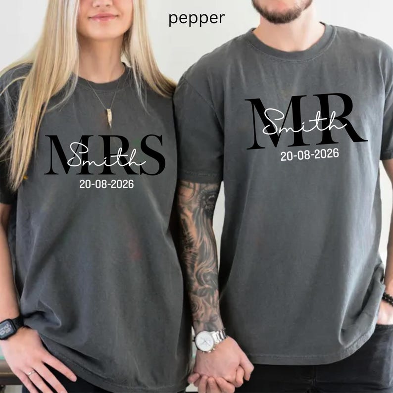 May include: Two pepper-colored t-shirts. One says "MRS Smith" and the other says "MR Smith" in black script. Both shirts have the date "20-08-2026" printed below the name. The shirts are likely for a couple.