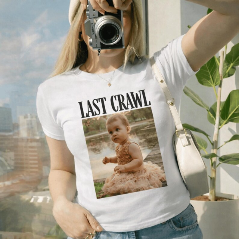 May include: White t-shirt featuring the text "LAST CRAWL" above a photograph of a baby in a peach dress. The image is set against a natural backdrop. The shirt is worn by a person holding a camera.
