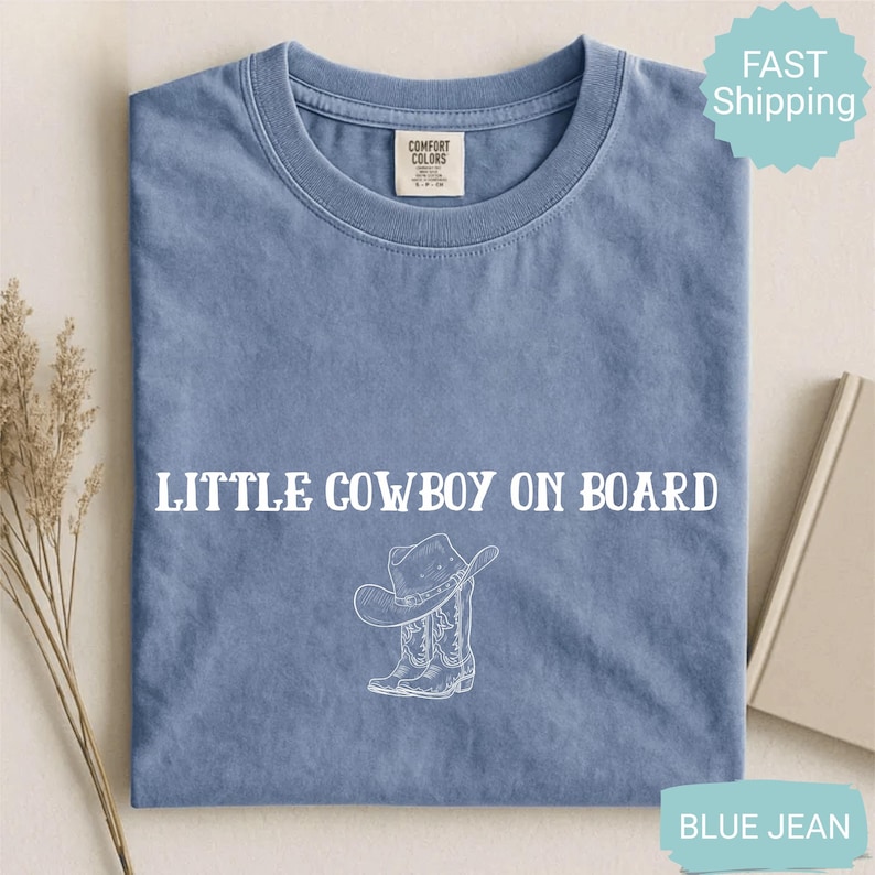 May include: A blue jean t-shirt with the text "LITTLE COWBOY ON BOARD" in white. Below the text is a white illustration of a cowboy hat and boots. The shirt has a crew neck and short sleeves.