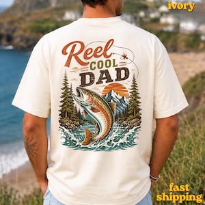 May include: Ivory-colored t-shirt with a graphic featuring a leaping trout, mountains, and the text "Reel Cool Dad." The design includes a fishing lure and a sunset. The shirt is a comfortable, casual wear.