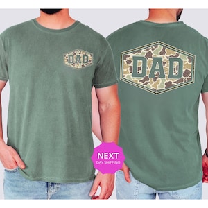 May include: Green t-shirt with a camouflage "DAD" design on the front and back. The text is in a hexagon shape. The shirt is a solid green color and has short sleeves.