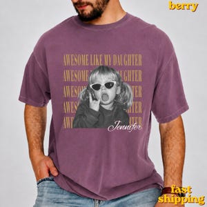 May include: A berry-coloured short-sleeve t-shirt featuring a black and white photograph of a child wearing sunglasses and sticking out their tongue. The text "AWESOME LIKE MY DAUGHTER" is repeated in gold, with the name "Jennifer" written below.