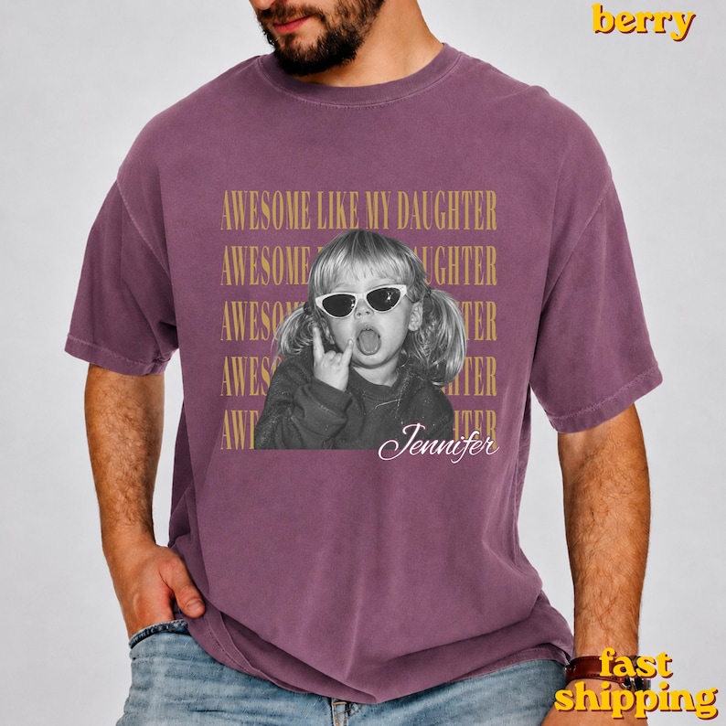 May include: A berry-coloured short-sleeve t-shirt featuring a black and white photograph of a child wearing sunglasses and sticking out their tongue. The text "AWESOME LIKE MY DAUGHTER" is repeated in gold, with the name "Jennifer" written below.