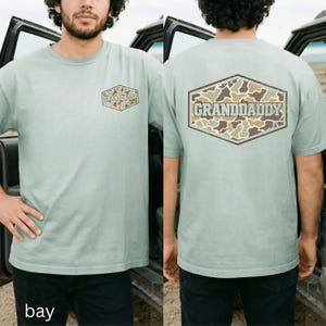 May include: A light green t-shirt with the word "GRANDADDY" printed on the front and back. The text is inside a brown and green camouflage design. The shirt is a short-sleeved crew neck.