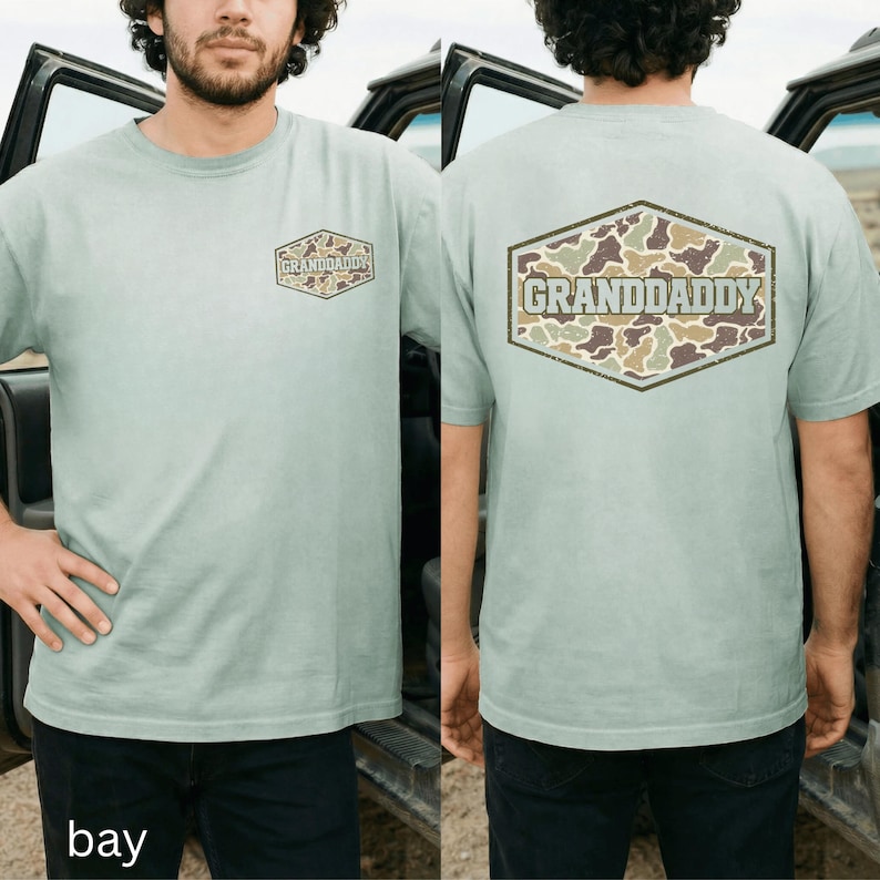 May include: A light green t-shirt with the word "GRANDADDY" printed on the front and back. The text is inside a brown and green camouflage design. The shirt is a short-sleeved crew neck.