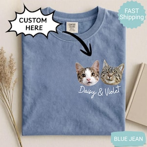 May include: Blue jean colored t-shirt with a custom cat face design. The shirt features two cat faces with the names "Daisy & Violet" printed below. The shirt has a crew neck and a tag that says "Comfort Colors".