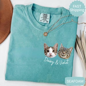 May include: Seafoam green t-shirt with two cat faces and the names "Daisy & Violet" in white script. The shirt has a crew neck and short sleeves. A gold necklace with a pendant is draped across the shirt.