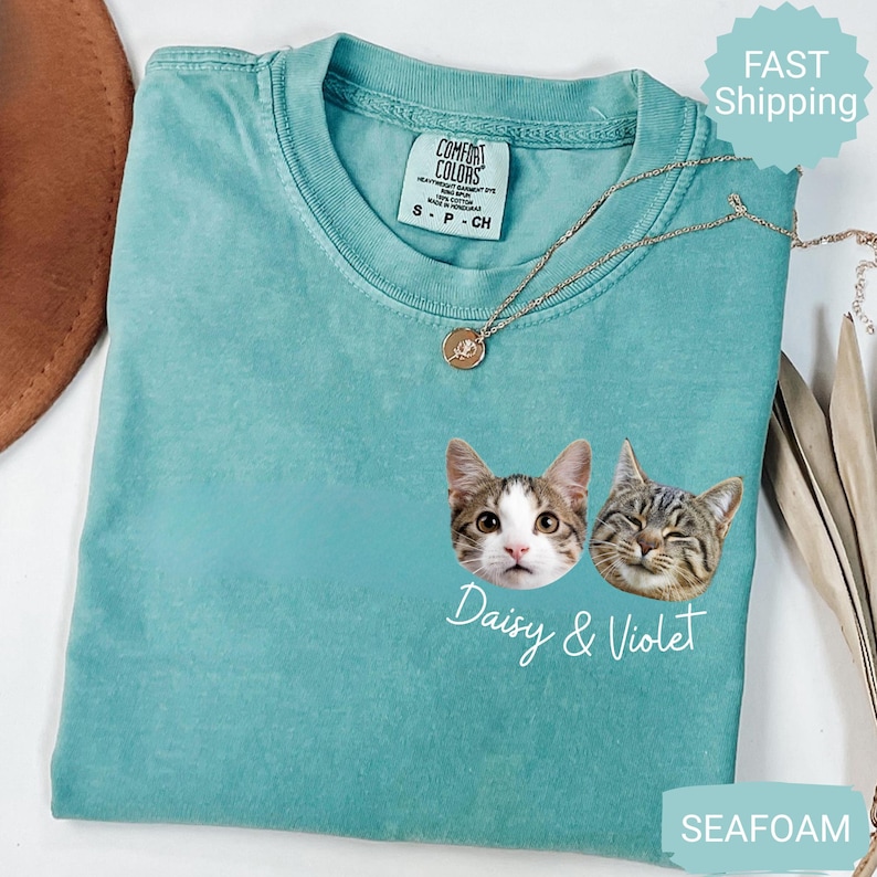 May include: Seafoam green t-shirt with two cat faces and the names "Daisy & Violet" in white script. The shirt has a crew neck and short sleeves. A gold necklace with a pendant is draped across the shirt.