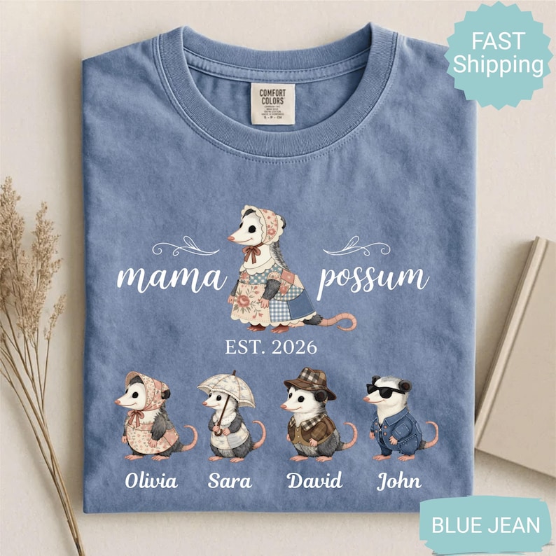 May include: Blue jean Comfort Colors t-shirt with a possum family graphic. The design features a mama possum and three possum babies, with the text "mama possum" and "EST. 2026". The names Olivia, Sara, David, and John are below the possum characters.