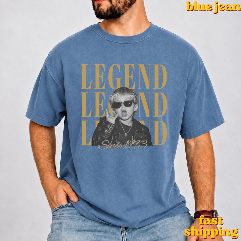 Puede incluir: Una camiseta azul de cuello redondo con una fotograf&iacute;a en blanco y negro de un ni&ntilde;o con gafas de sol y chaqueta de cuero. El texto LEGEND aparece en letras doradas grandes arriba y abajo, con la frase Since 1993 en letra cursiva dorada.