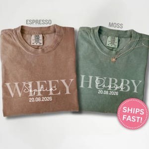 May include: Two t-shirts, one espresso-colored and one moss-colored, are displayed. The espresso shirt says "Wifey Sophia 20.08.2026" and the moss shirt says "Hubby David 20.08.2026". A pink sticker with "SHIPS FAST!" is visible.