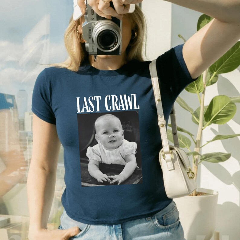 May include: A navy blue t-shirt featuring the words "LAST CRAWL" above a black and white image of a baby. The baby is smiling and wearing a white dress. The shirt is being worn by a person.