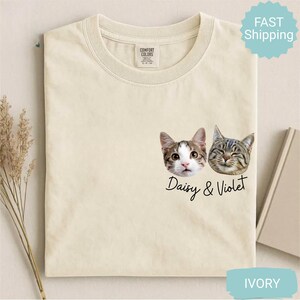 May include: Ivory-colored t-shirt featuring two cat faces, one with wide eyes and the other with closed eyes. The text "Daisy & Violet" is printed below the cat faces. The shirt has a crew neck and a tag that says "Comfort Colors".
