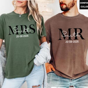 May include: Two short-sleeved t-shirts, one olive green and one brown. The olive green shirt says "MRS Smith 20-08-2026" in black script. The brown shirt says "MR Smith 20-08-2026" in black script. Both shirts are for a wedding.