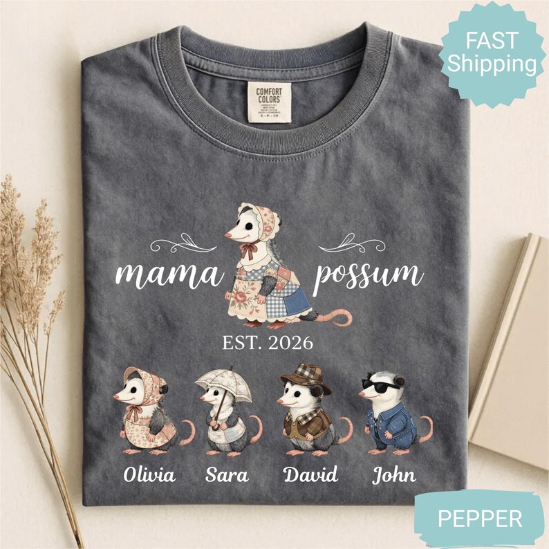 May include: Dark gray Comfort Colors t-shirt featuring a "Mama Possum" design with a possum wearing a bonnet and patchwork dress. Below are illustrations of possums named Olivia, Sara, David, and John. The text "EST. 2026" is also present.