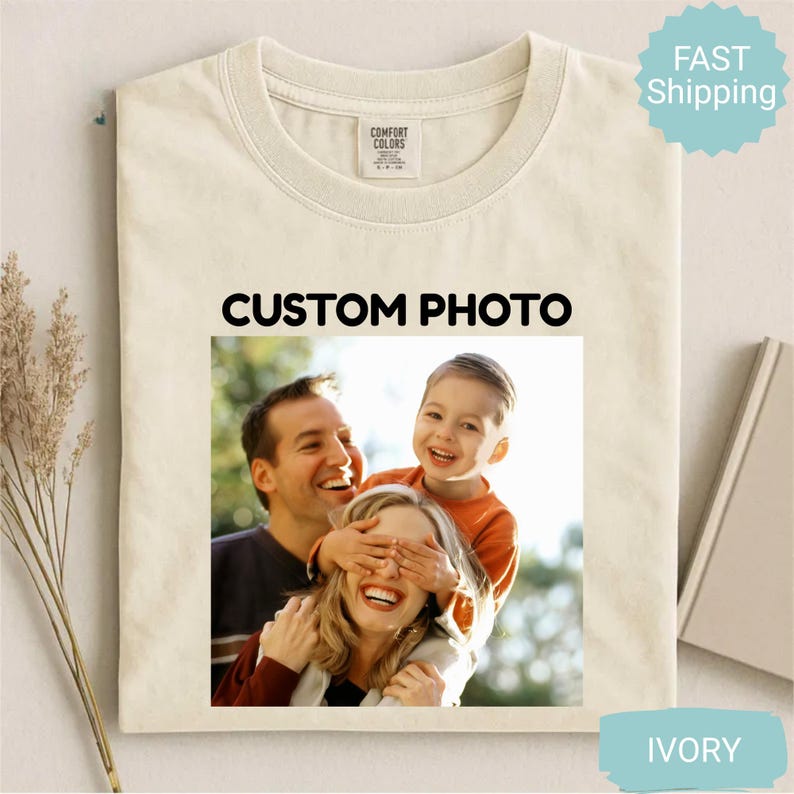 May include: Ivory-colored t-shirt with a family photo. The photo features a smiling family of three, with the text "CUSTOM PHOTO" above the image. The shirt has a crew neck and a tag that says "COMFORT COLORS".