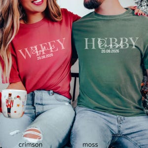 May include: Crimson and moss green t-shirts with white text. The crimson shirt says "Wifey Sophia 20.08.2026". The moss green shirt says "Hubby David 20.08.2026". Both shirts are short-sleeved and worn by people.