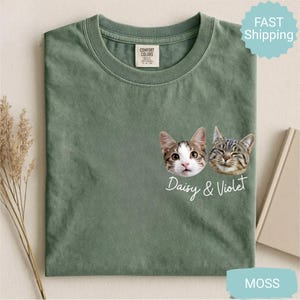 May include: Moss green t-shirt featuring two cat faces with the text "Daisy & Violet." The shirt has a crew neck and a small tag on the upper left. The shirt is made of a soft material.