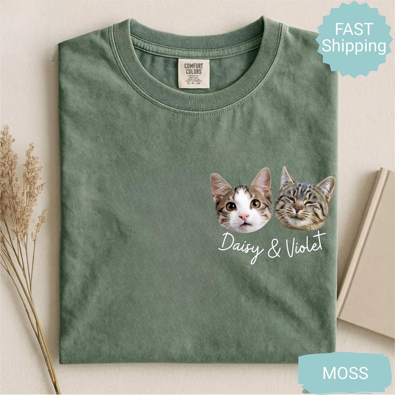 May include: Moss green t-shirt featuring two cat faces with the text "Daisy & Violet." The shirt has a crew neck and a small tag on the upper left. The shirt is made of a soft material.