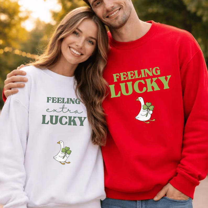 May include: White and red sweatshirts with a St. Patrick's Day theme. The white sweatshirt has the text "Feeling extra LUCKY" and a goose holding a four-leaf clover. The red sweatshirt has the text "FEELING LUCKY" and a goose holding a four-leaf clover.