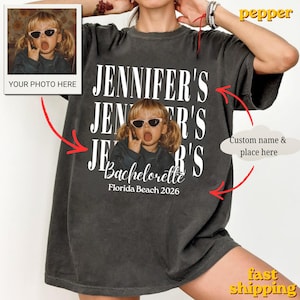 Puede incluir: Una camiseta gris carbón de manga corta con un estampado de texto blanco repetido del nombre Jennifer's. En el centro hay un gráfico de un niño con gafas de sol haciendo un gesto con la mano, con el texto Bachelorette Florida Beach 2026.