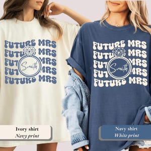 K&ouml;nnte beinhalten: Zwei T-Shirts, eines in Elfenbein und eines in Marineblau, zeigen den Text "FUTURE MRS" in einem sich wiederholenden Muster. Eine Diamantring-Grafik umgibt den Namen "Smith". Das elfenbeinfarbene Shirt hat einen marineblauen Aufdruck, das marineblaue Shirt einen wei&szlig;en Aufdruck.