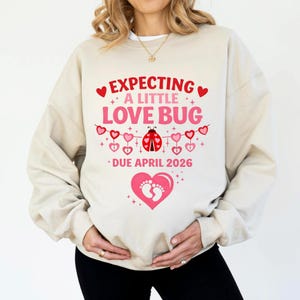 Op de afbeelding: Beige sweatshirt met de tekst "EXPECTING A LITTLE LOVE BUG" in rood en roze. Een lieveheersbeestje-afbeelding is gecentreerd onder de tekst, met een banner van harten. De woorden "DUE APRIL 2026" staan eronder. Een roze hart met voetafdrukken staat onderaan.